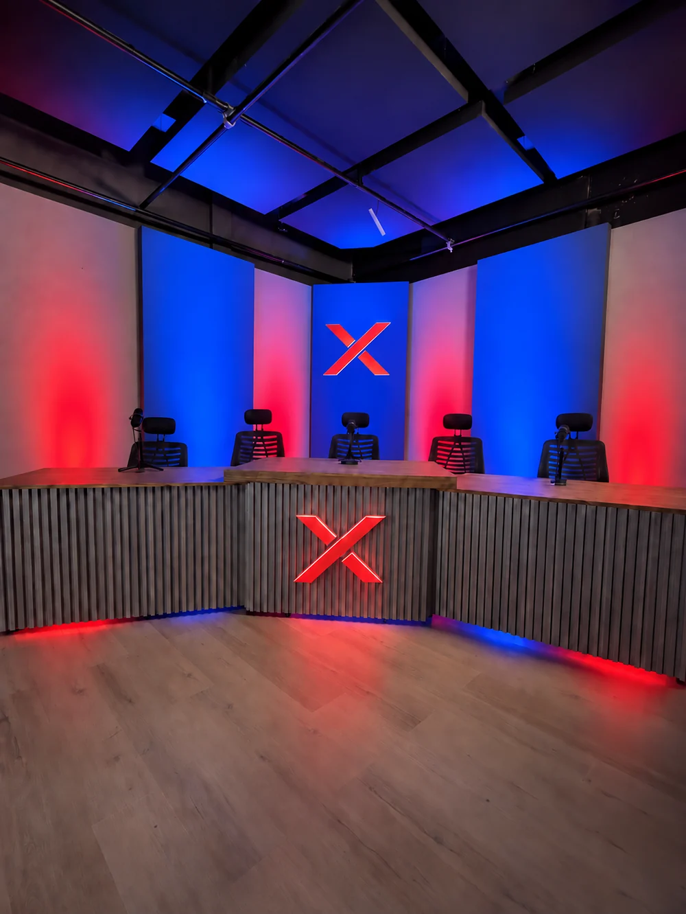 Set de streaming de NEXO Studios con cinco puestos con micrófono, mostrador de madera y chapa acanalada, paneles azules y rojos y el logo X de NEXO