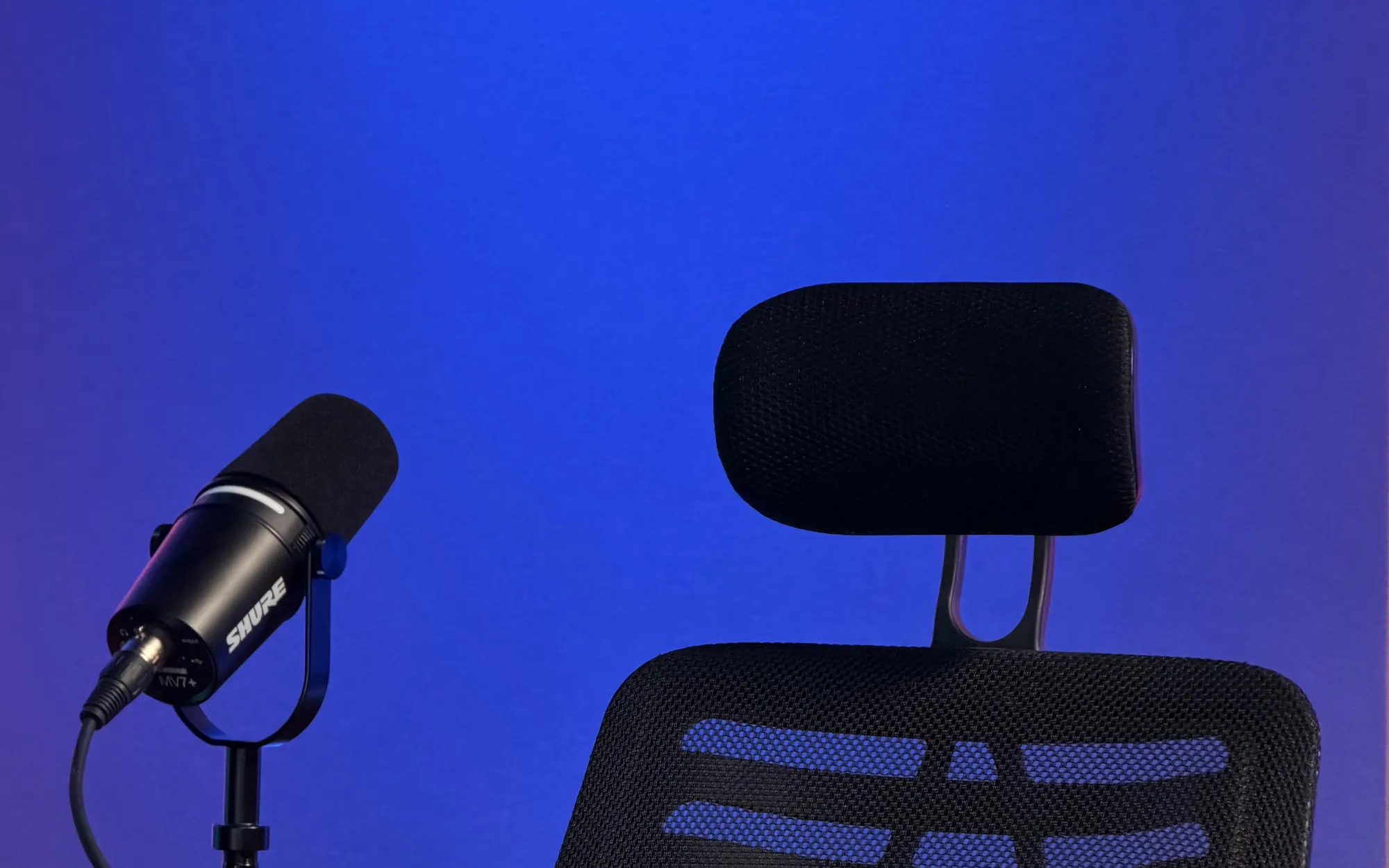 Primer plano de un micrófono Shure MV7+ y una silla de conducción sobre fondo azul
